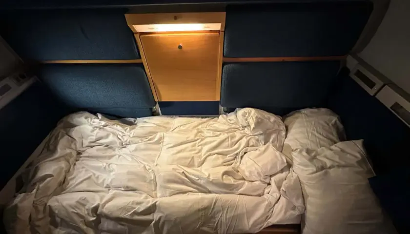 Ett bäddat nattågskupé med vita lakan och kuddar. En liten trähylla med lampa finns ovanför sängen.