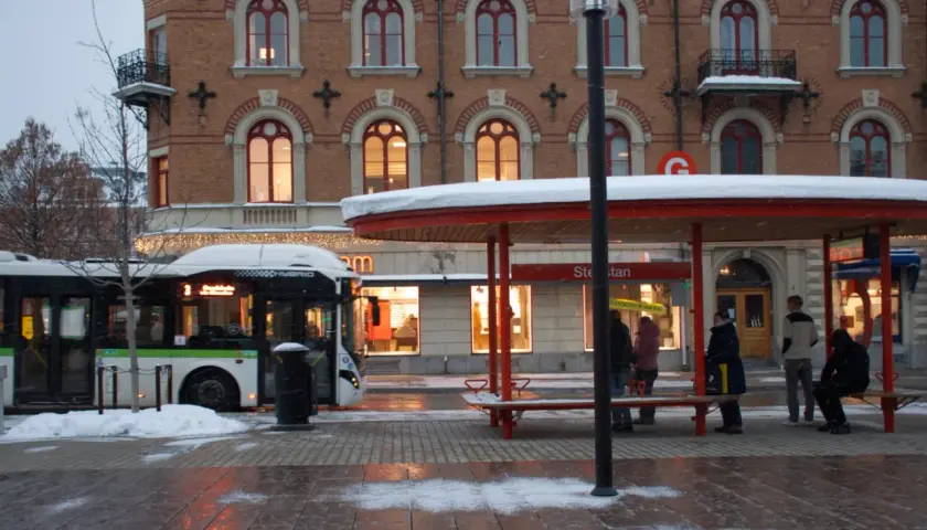 Snö faller vid en busshållplats där folk väntar. En buss med destination "tåg till Sundsvall" står parkerad.