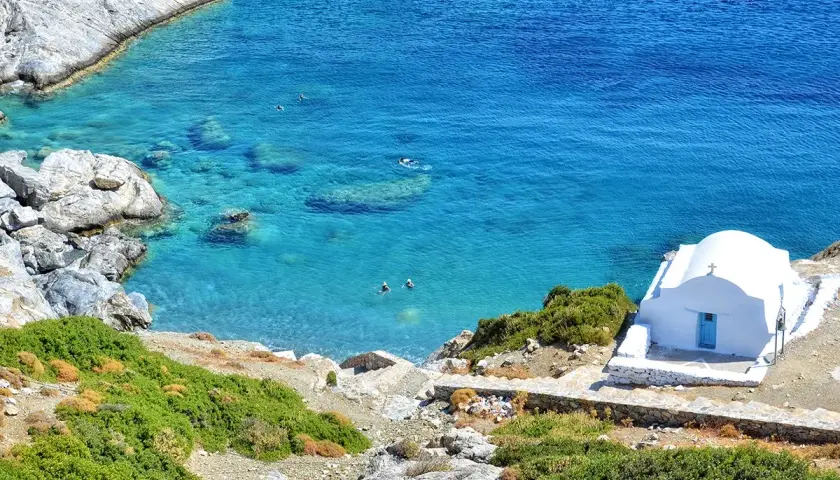 En vit kyrka med blå dörr vid en turkos vik på Amorgos, Grekland. Några badar i det klara vattnet.