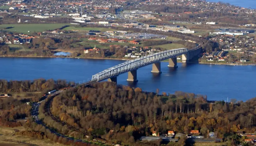 Lilla Bältbron sträcker sig över det blå vattnet, omgiven av höstfärgad skog och en stad i bakgrunden.
