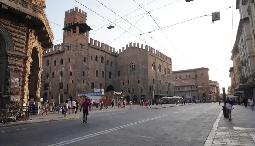 En solig dag i Bologna, Italien. En stor medeltida byggnad med torn dominerar torget, där människor promenerar och sitter vid uteserveringar.