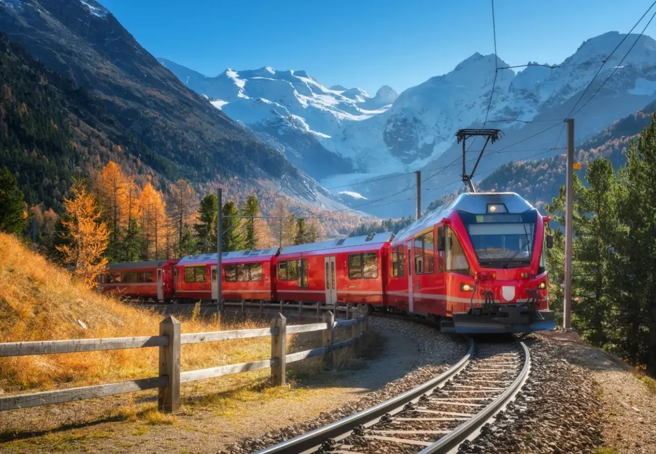 Rött tåg på Bernina Express-linjen, perfekt för Interrail, slingrar sig genom en höstfärgad dal med snöklädda berg i bakgrunden.