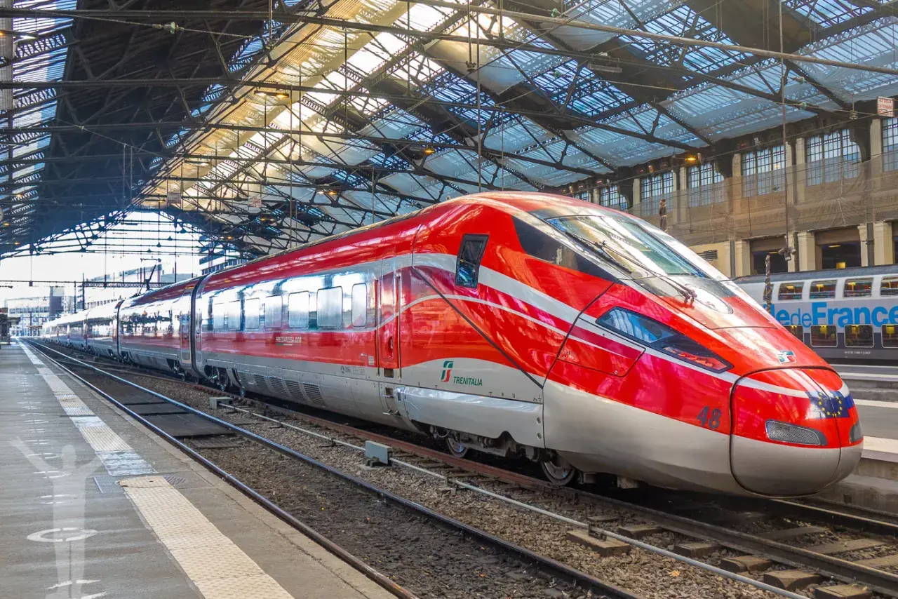 Ett rött snabbtåg i Frankrike, med namnet "Frecciarossa 1000", står på en station under ett glastak.