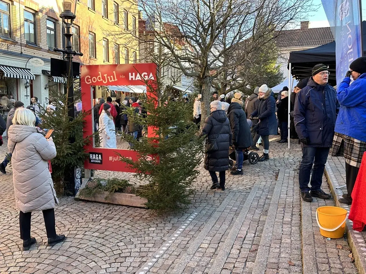 Människor njuter av en livlig julmarknad i Haga. En röd ram med texten "God Jul från HAGA" inbjuder till foton bland granar och stånd.