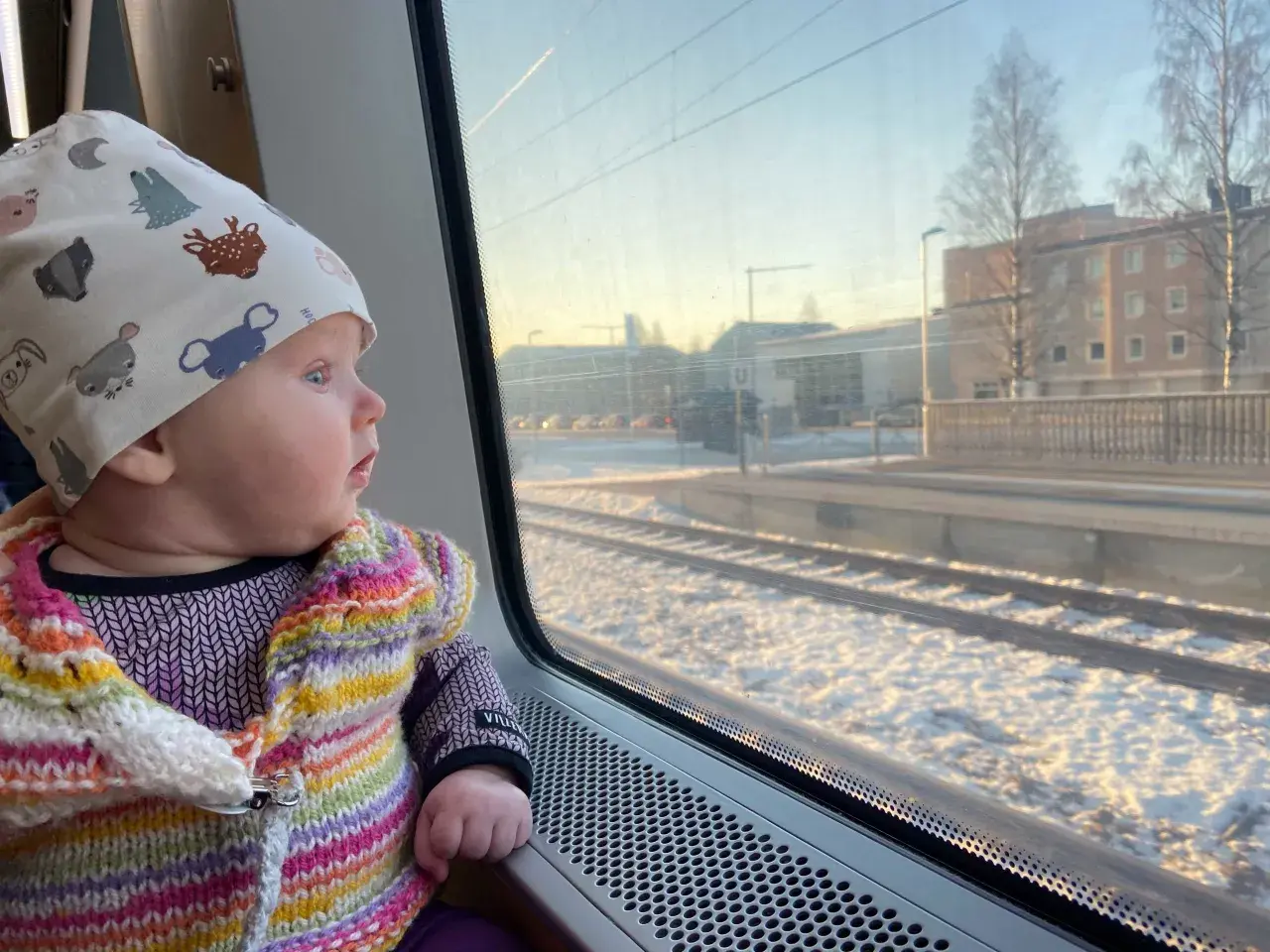 Litet barn i färgglad kofta och mössa med djurmotiv tittar ut genom tågfönstret. Snö på marken och byggnader syns utanför. Kanske är det ett nattåg till Bryssel?