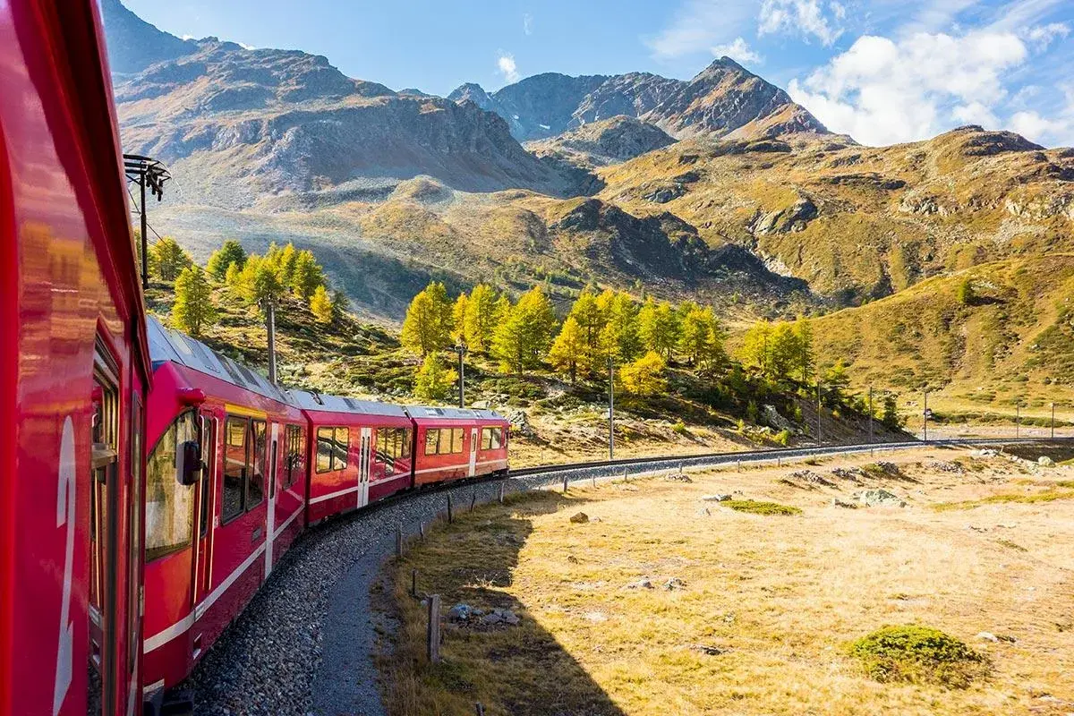 Rött tåg slingrar sig genom ett bergigt landskap med höstfärger. Ett perfekt sätt att uppleva Europa, funderar du på interrail pris?