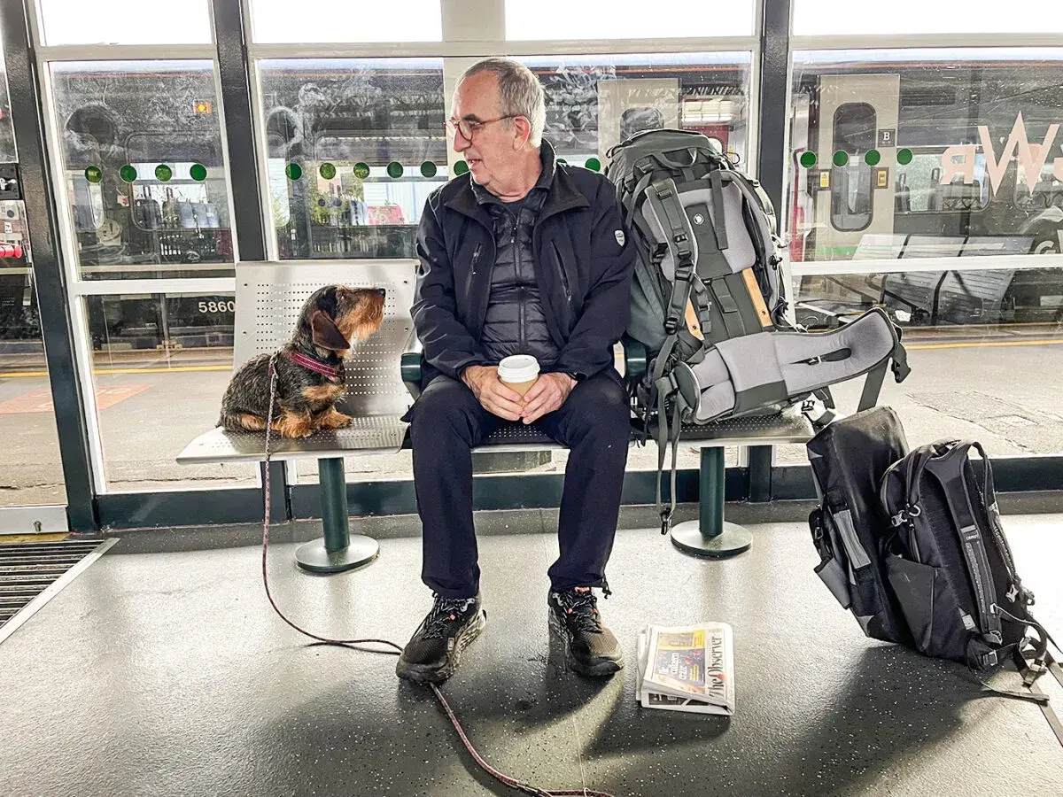 Man och hund väntar på tåget för att tågluffa Skottland. Stor ryggsäck och tidning redo för äventyret.