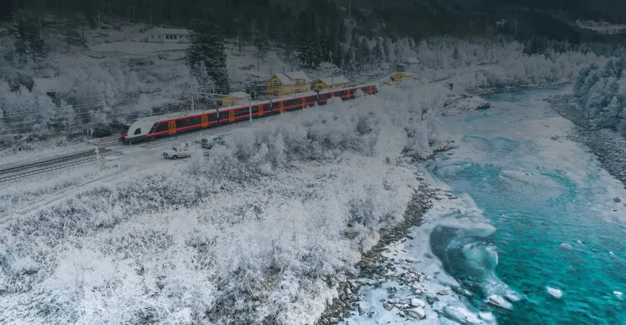 Ett snötäckt landskap med ett rött och vitt tåg som färdas längs spåret. En flod med iskanter flyter bredvid.