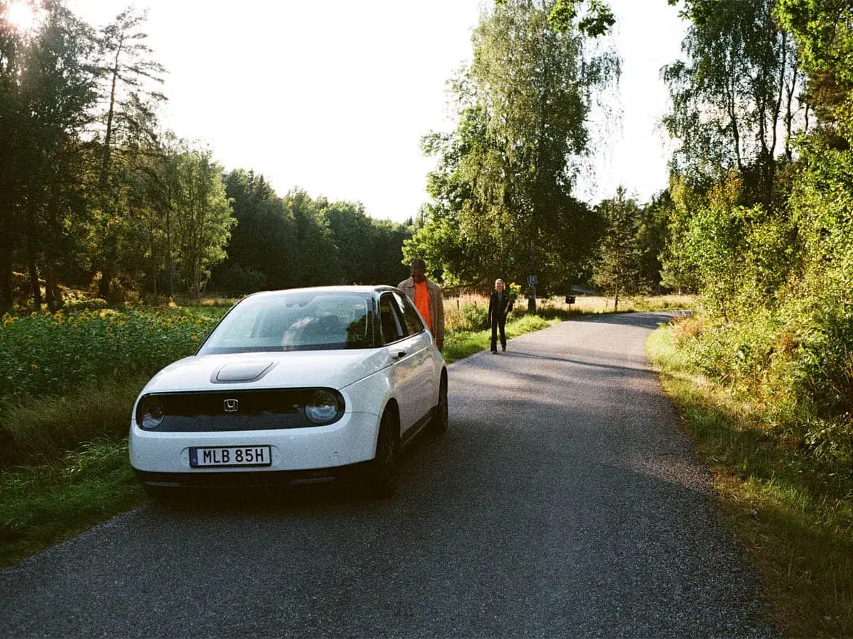 Vit elbil parkerad vid en skogsväg, redo för resan från Stockholm till Oslo. Två personer står vid sidan av vägen.