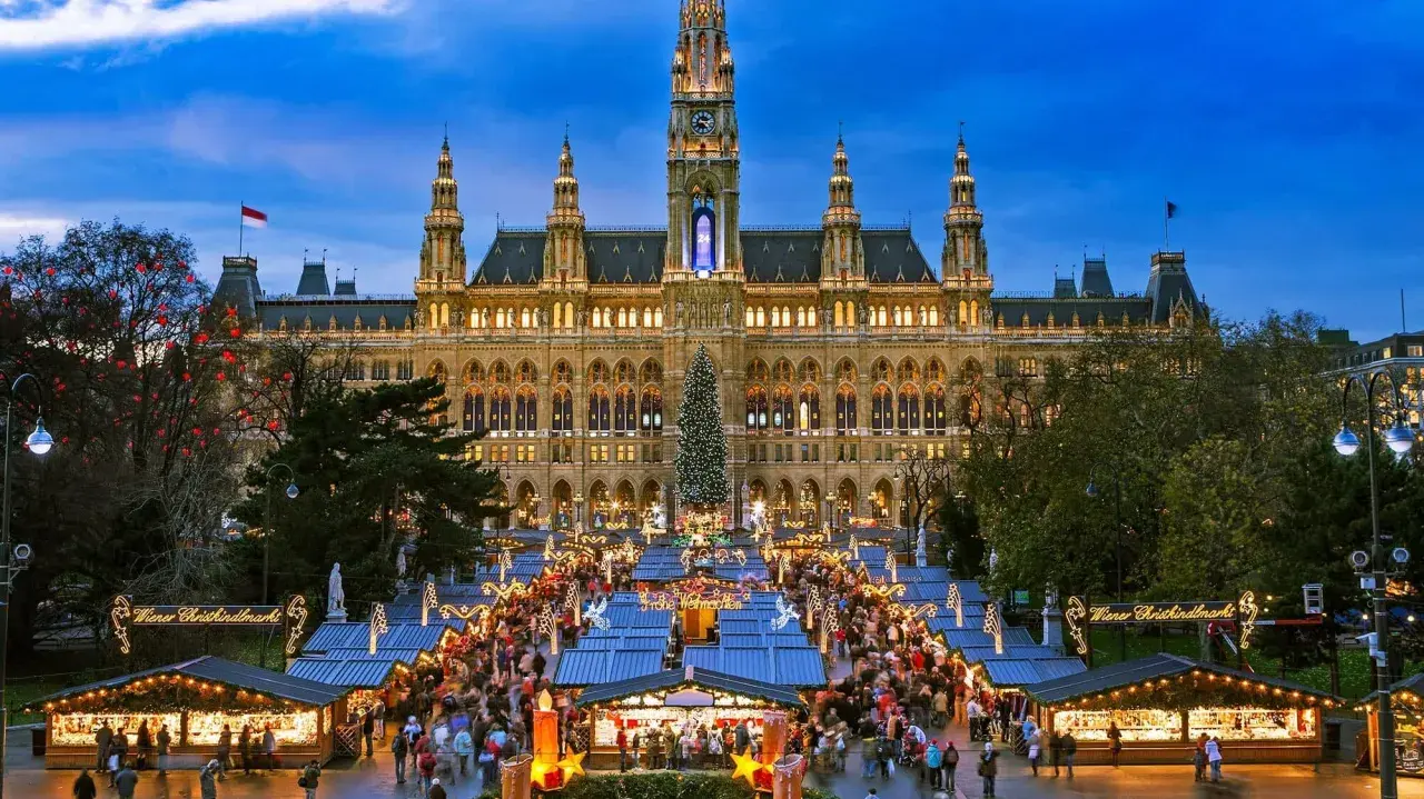 En livlig julmarknad i Europa framför ett upplyst stadshus. Träd med röda dekorationer och en stor julgran bidrar till den festliga stämningen.