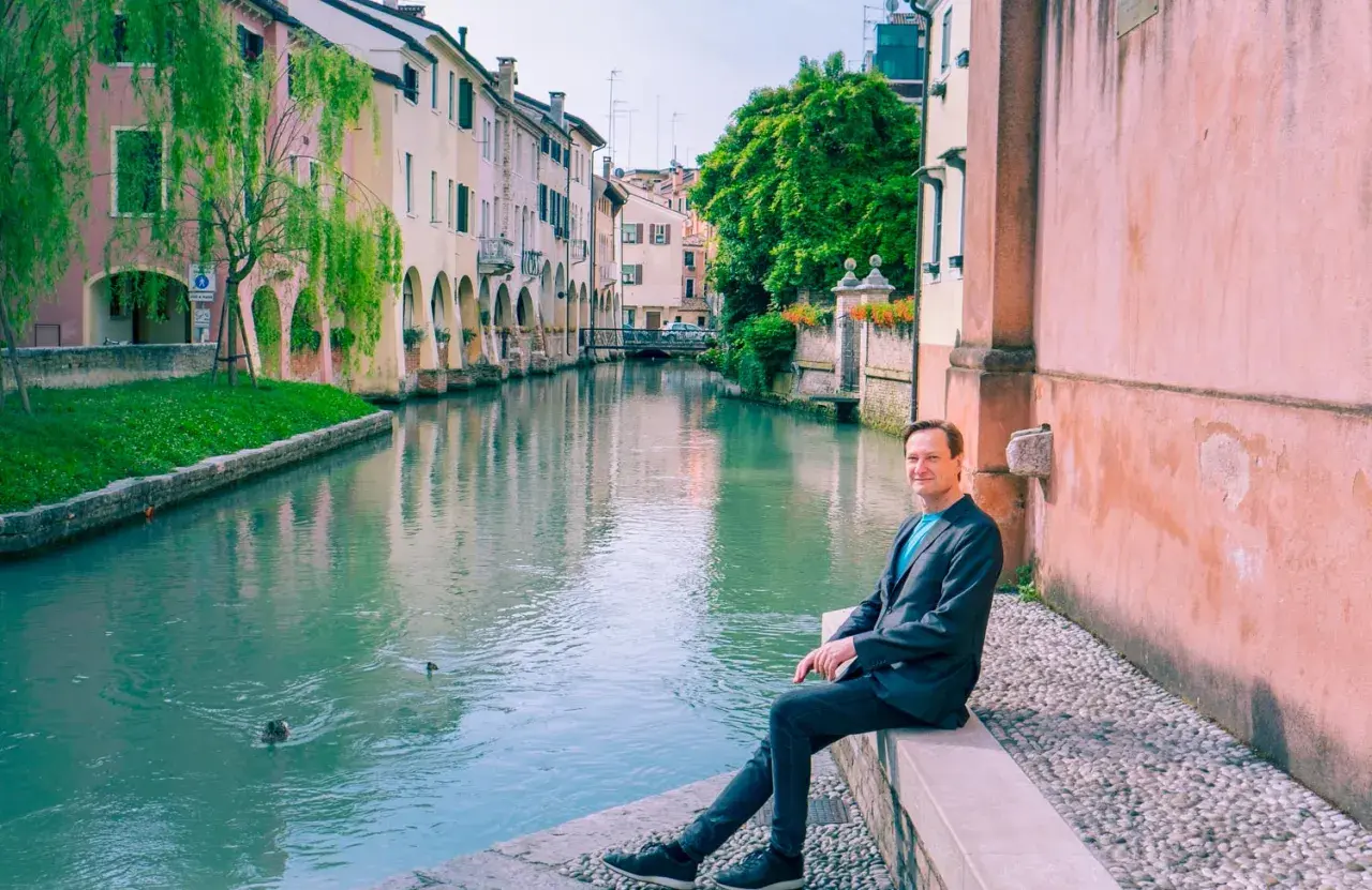 Man sitter vid en kanal i Treviso, Italien. Vatten reflekterar färgglada hus och gröna träd.