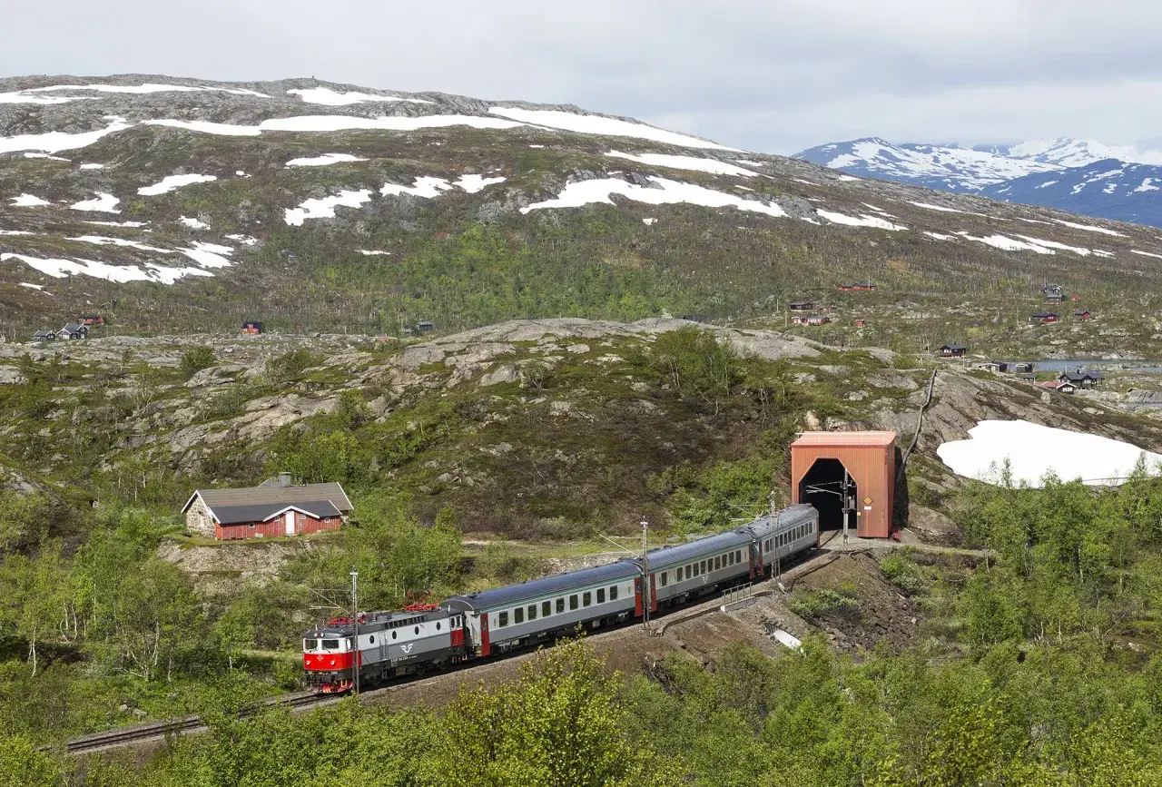 Tåg på väg mot tunneln, en del av sträckan Kiruna Narvik, med snö på bergssluttningarna och små röda hus.