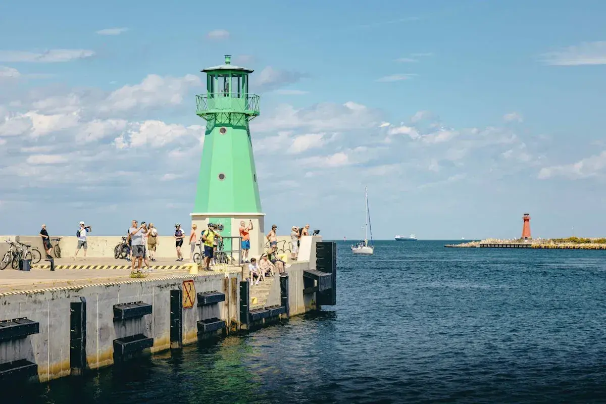 Folk njuter av solen vid en grön fyr, en vy som påminner om Karlskrona eller Gdansk. En segelbåt glider förbi i det blå vattnet.