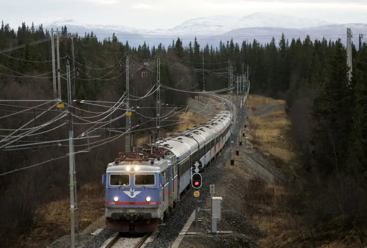 Ett SJ-tåg med nummer 1343 på väg mot Trondheim kurvar genom en skog med snöklädda berg i bakgrunden.