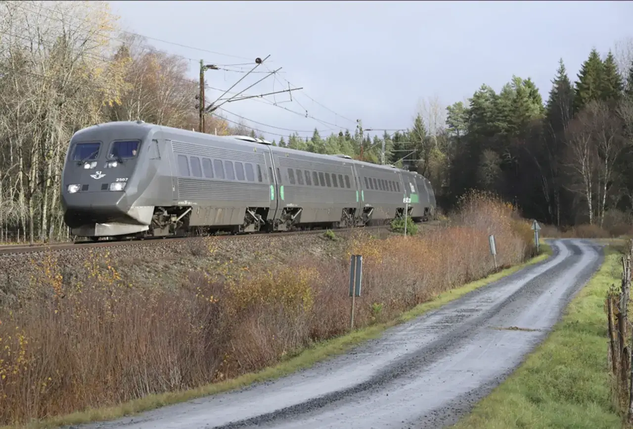 Grått SJ-tåg på väg mot Stockholm, passerar en slingrande grusväg och höstfärgad natur.