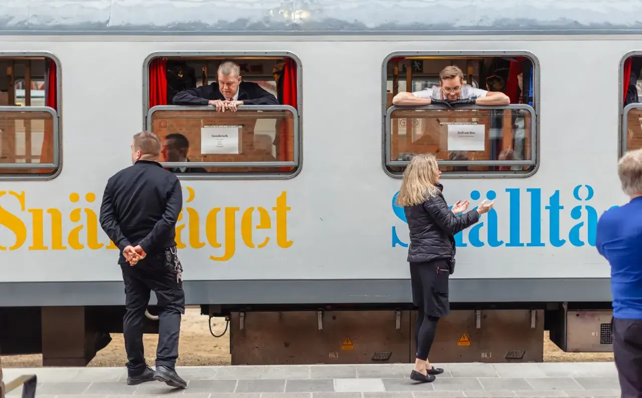 Män tittar ut genom fönstret på ett tåg med texten "Snälltåget" och destinationen Innsbruck. En kvinna gestikulerar mot tåget.