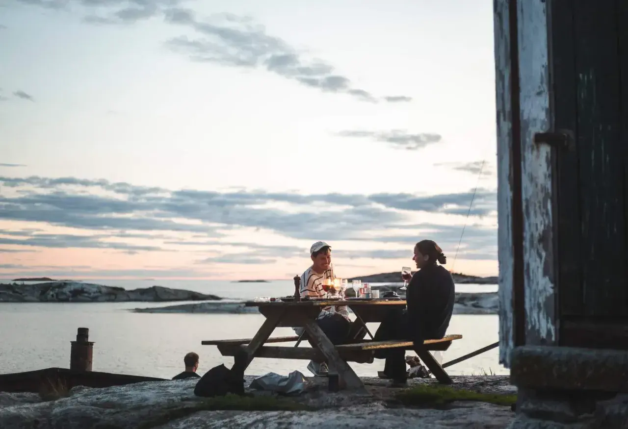 Två personer njuter av en middag vid ett picknickbord vid vattnet. En perfekt hemester i Stockholms skärgård.