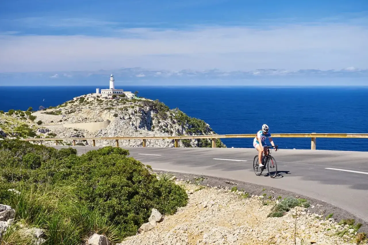En cyklist njuter av en cykelsemester i Europa, med en fyr på en klippa vid det blå havet i bakgrunden.