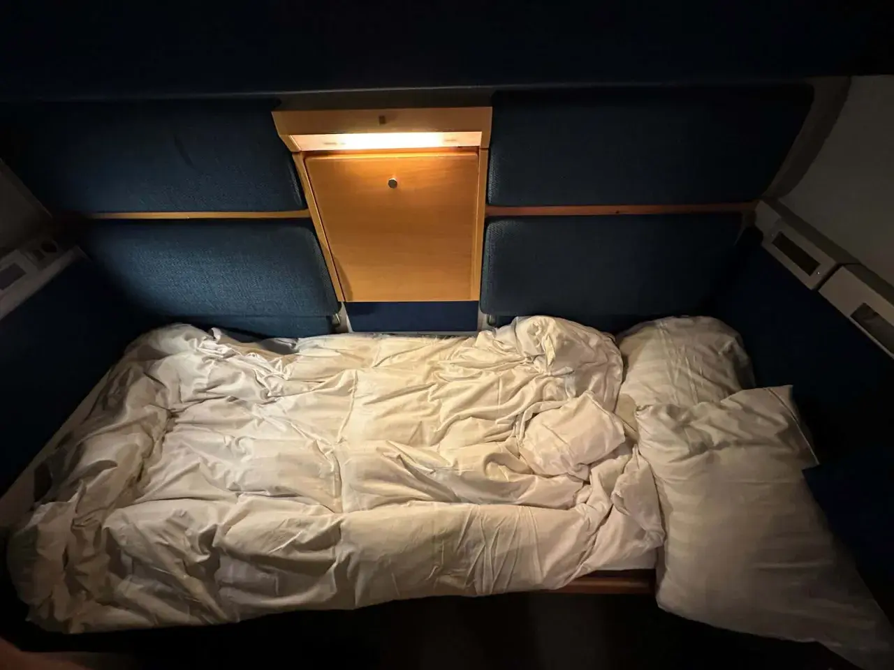 Ett bäddat nattågskupé med vita lakan och kuddar. En liten trähylla med lampa finns ovanför sängen.