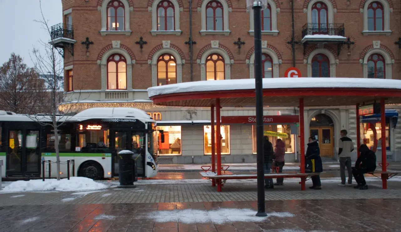 Snö faller vid en busshållplats där folk väntar. En buss med destination "tåg till Sundsvall" står parkerad.