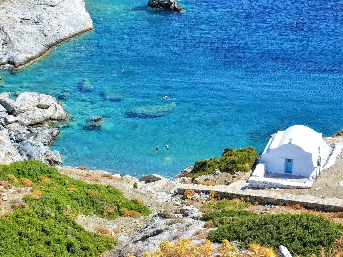 En vit kyrka med blå dörr vid en turkos vik på Amorgos, Grekland. Några badar i det klara vattnet.