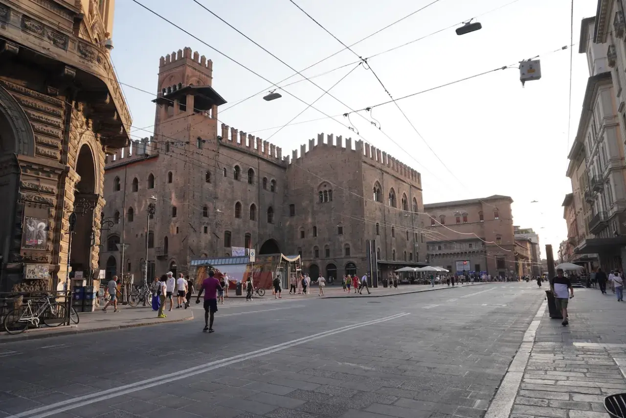 En solig dag i Bologna, Italien. En stor medeltida byggnad med torn dominerar torget, där människor promenerar och sitter vid uteserveringar.