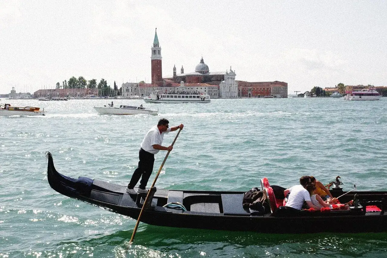 En gondoljär styr sin gondol med passagerare genom Venedigs kanaler, med kyrkor och palats i bakgrunden. En perfekt resa med tåg till Venedig.