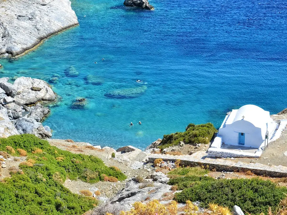 En vit kyrka med blå dörr vid en turkos vik på Amorgos, Grekland. Några badar i det klara vattnet.