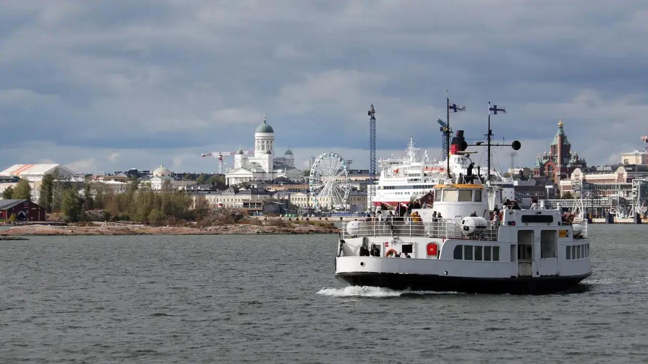 En f&auml;rja med passagerare n&auml;rmar sig Helsingfors skyline, med katedralen och ett pariserhjul synliga. Perfekt f&ouml;r en weekend Helsingfors.