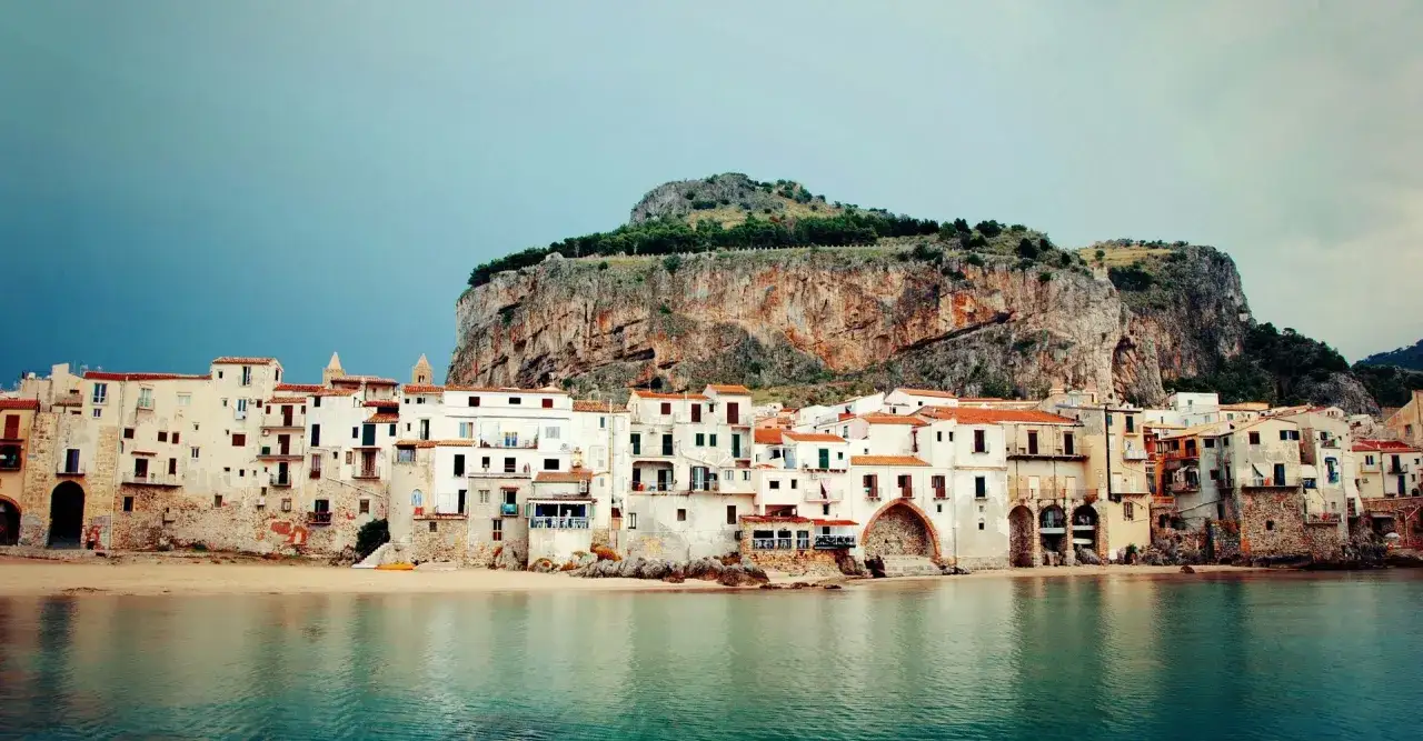 En rad vita hus med orange tak vid en strand, bakom en klippa. Ett av många mysiga resmål i Italien.