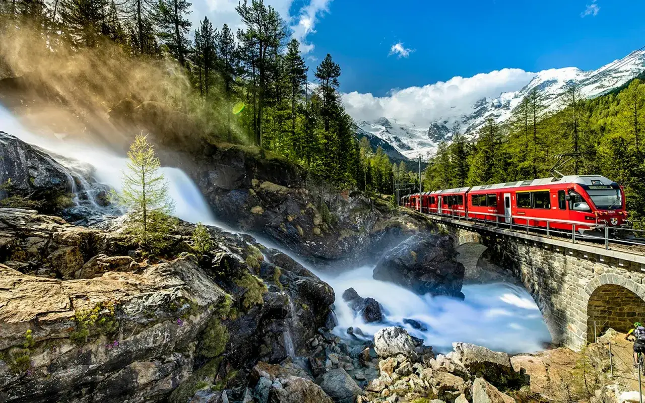 Bernina Express på en bro över ett vattenfall, med snöklädda berg i bakgrunden. En dröm för Interrail-resenärer.