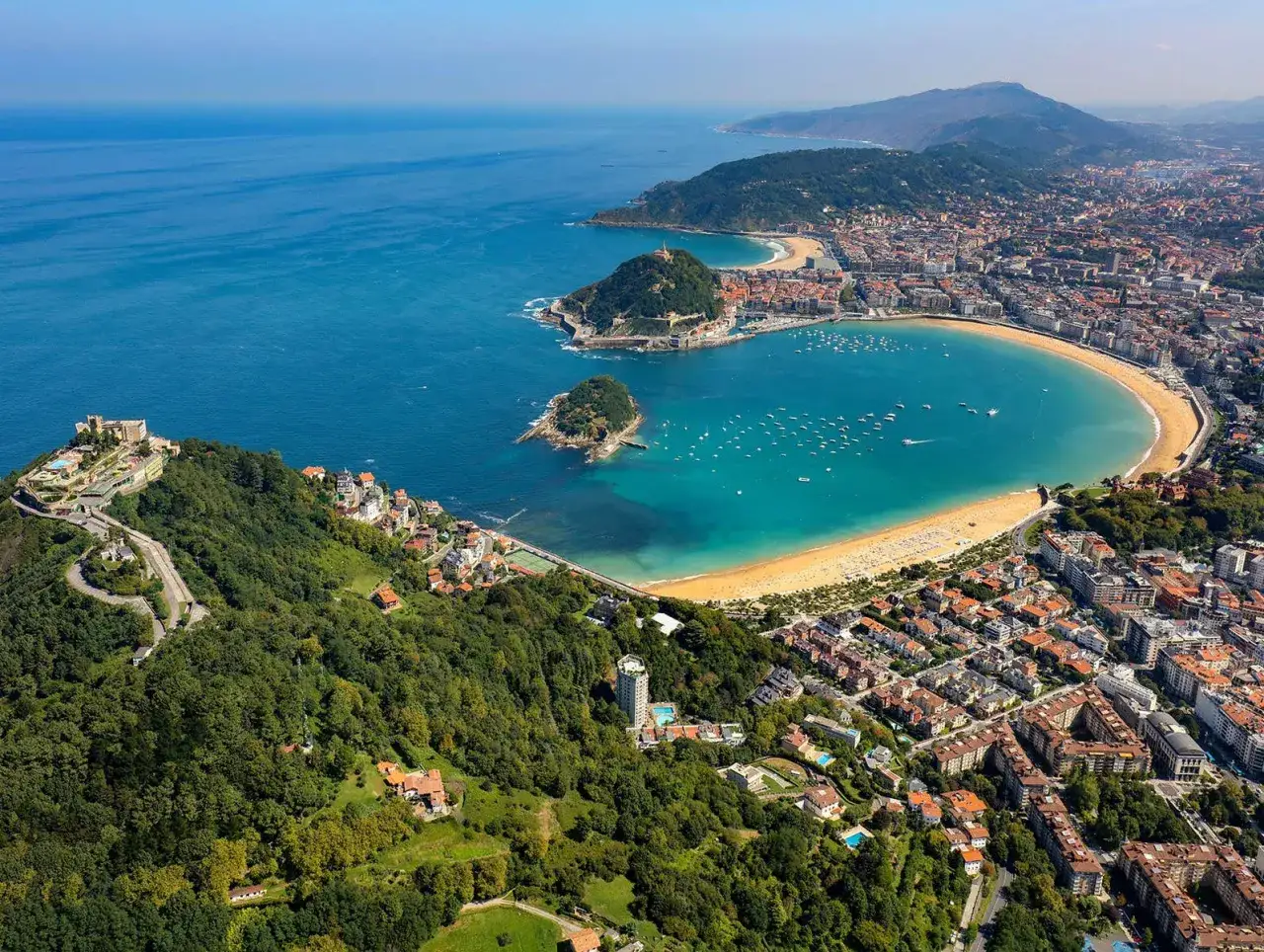 Luftvy över San Sebastián med en grönskande kulle med ett slott, en gyllene strand och turkost vatten fyllt av båtar.
