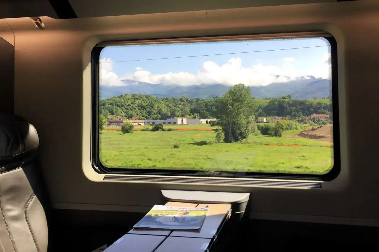 Vy från tågfönstret på en grönskande landsbygd med berg i bakgrunden, en försmak av nattåg till Italien.