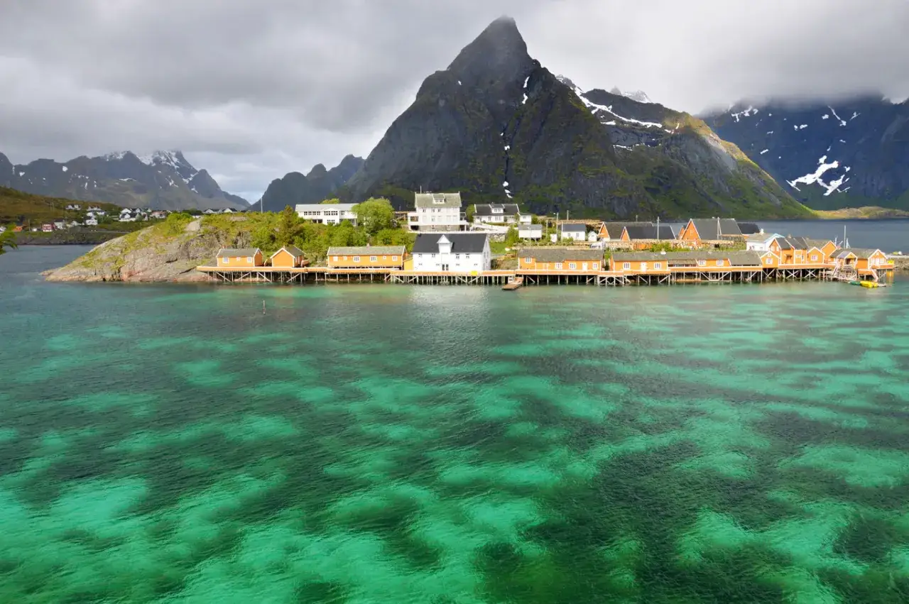 En dröm att bila till Lofoten! Röda stugor på pålar vid turkost vatten, med dramatiska berg bakom.