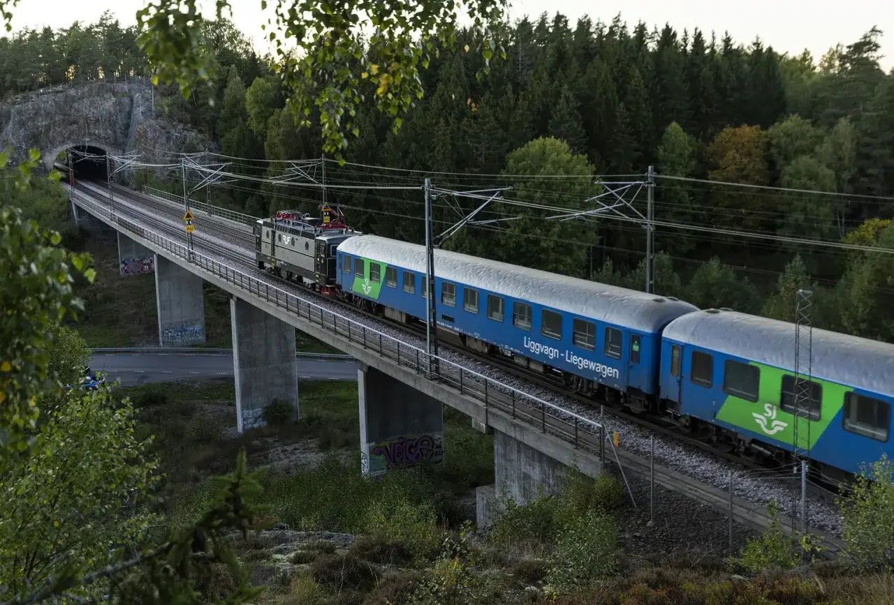 Ett SJ-tåg med blåa vagnar passerar över en bro genom en skogsmiljö. Tåget närmar sig en tunnel i en bergssida.