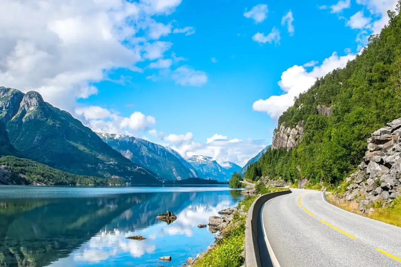 En vacker väg slingrar sig längs en spegelblank sjö i Norge. Berg täckta av grönska reser sig majestätiskt mot en klarblå himmel med vita moln. En dröm för alla som älskar att bila i Norge.
