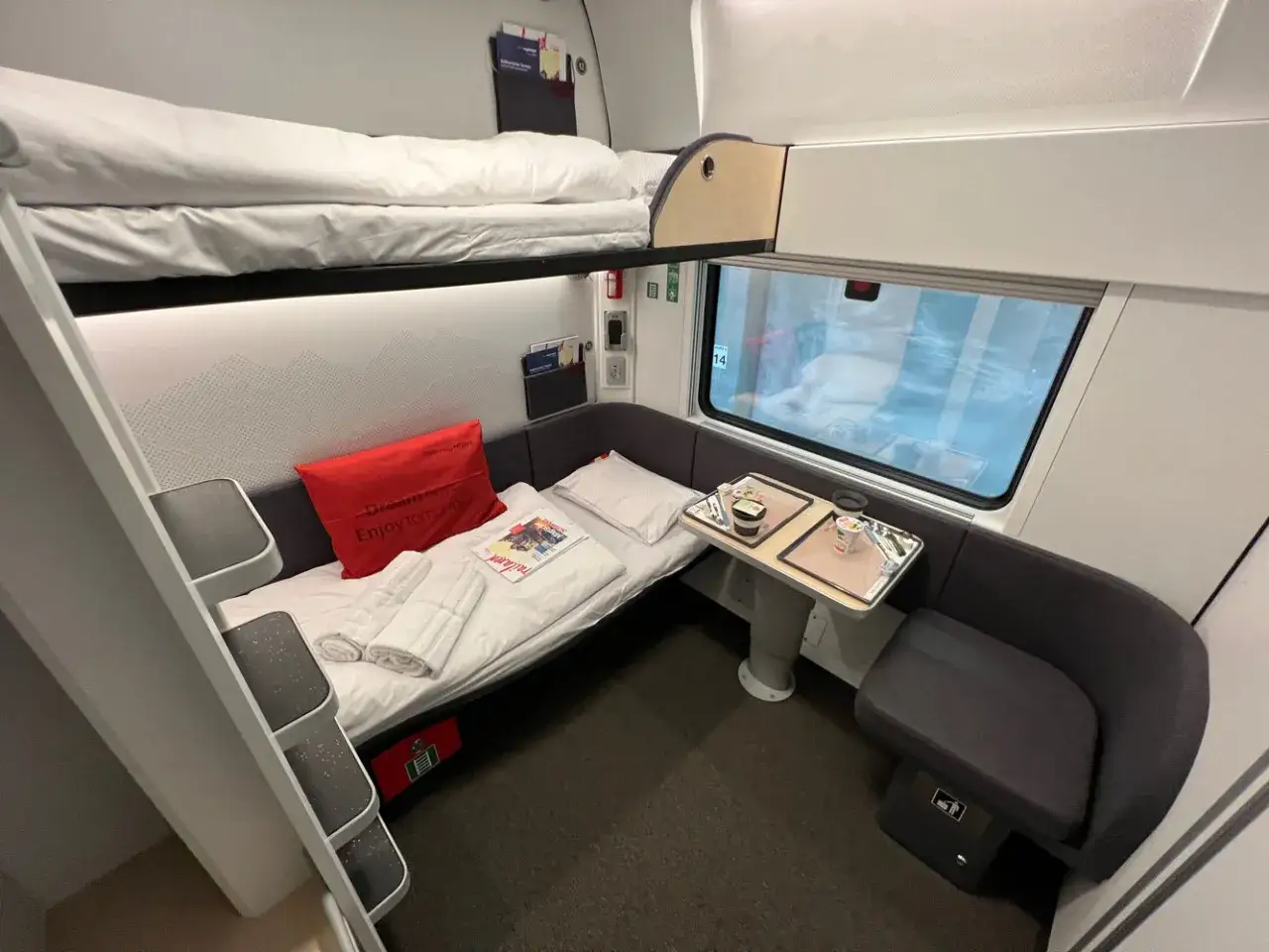 En sovhytt på ett Nightjet-tåg med våningssängar, en bekväm sittgrupp och ett bord med frukost.
