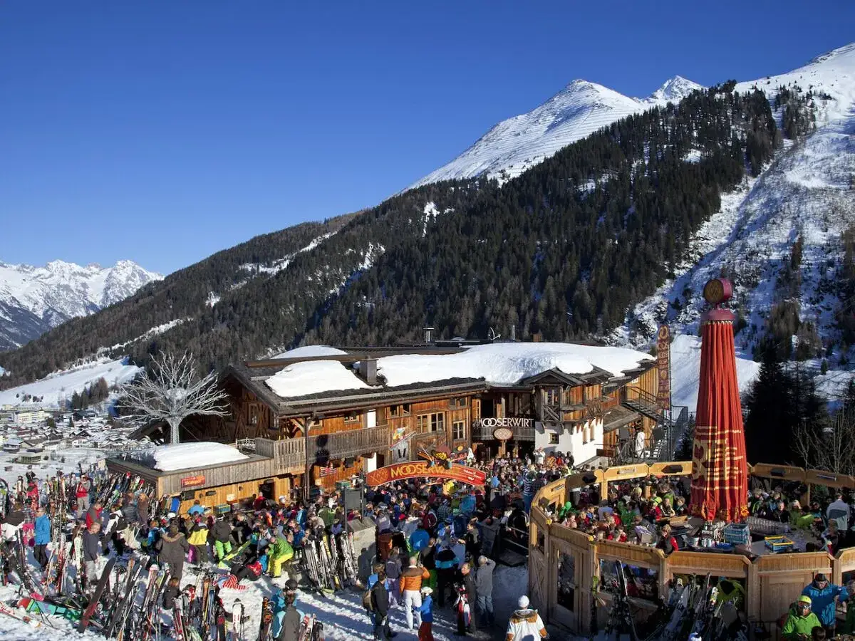 En livlig afterski vid Mooserwirt, en populär destination för alla som tar tåg till St. Anton. Skidåkare minglar i snön bland berg och träd.