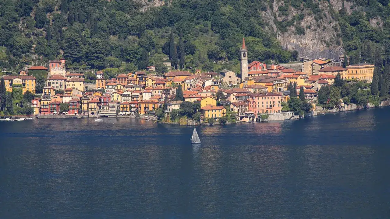 Färgglada, mysiga byar i norra Italien längs en sjö, med en segelbåt i förgrunden och gröna berg i bakgrunden.