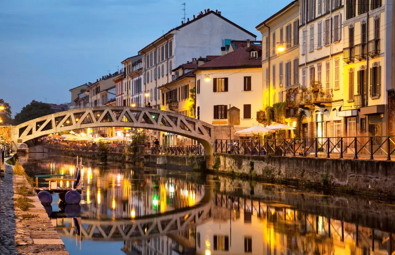 En charmig bro över en kanal i Milano, med upplysta byggnader och reflektioner i vattnet. Perfekt för en milano reseguide.