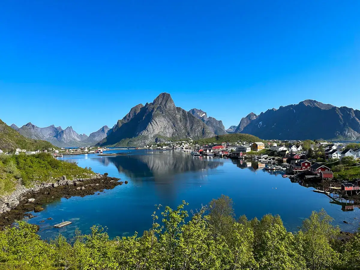 En klarblå himmel speglar sig i det lugna vattnet vid en pittoresk by. Majestätiska berg reser sig bakom de röda husen, en idyllisk vy av Lofoten sommar.
