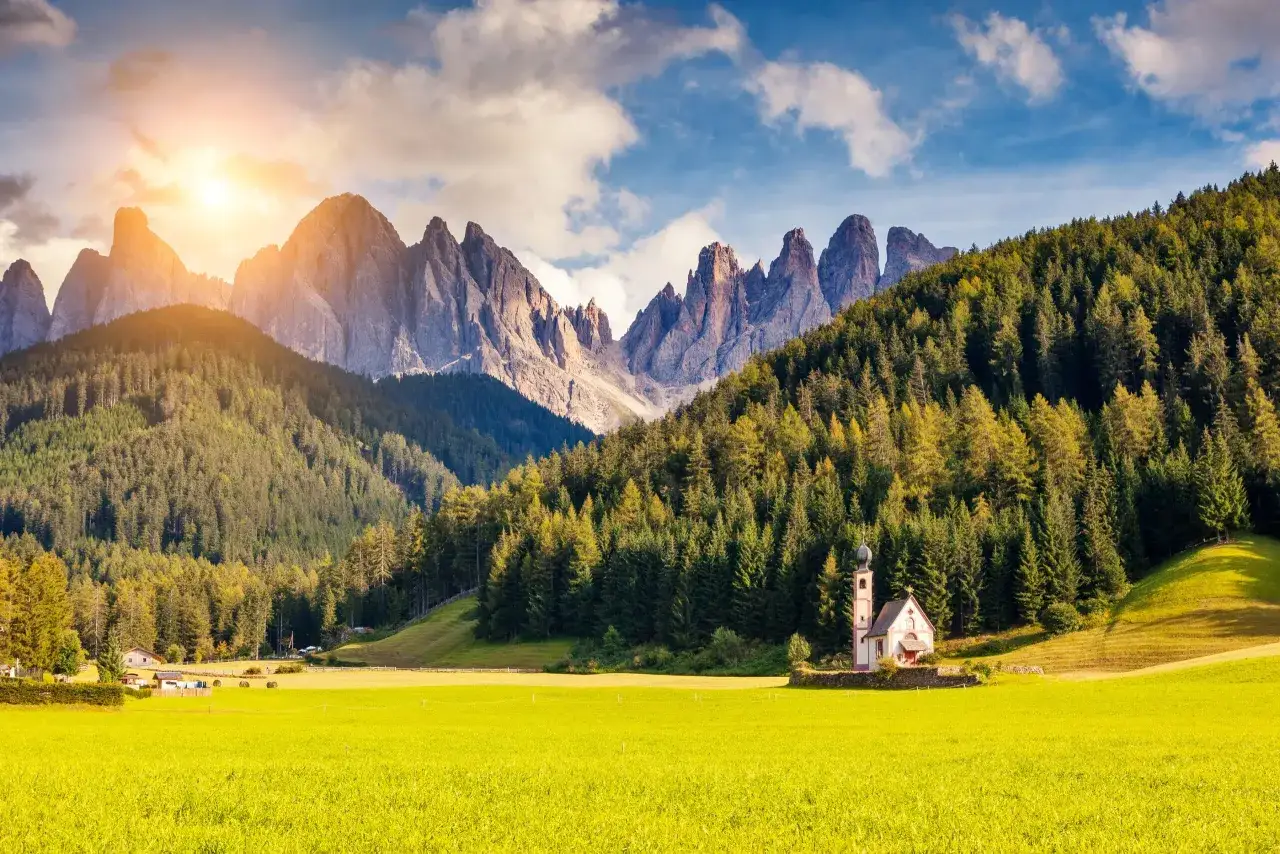 En solig sommardag i Dolomiterna. En liten kyrka står på en grön äng framför en tät granskog och dramatiska bergstoppar.
