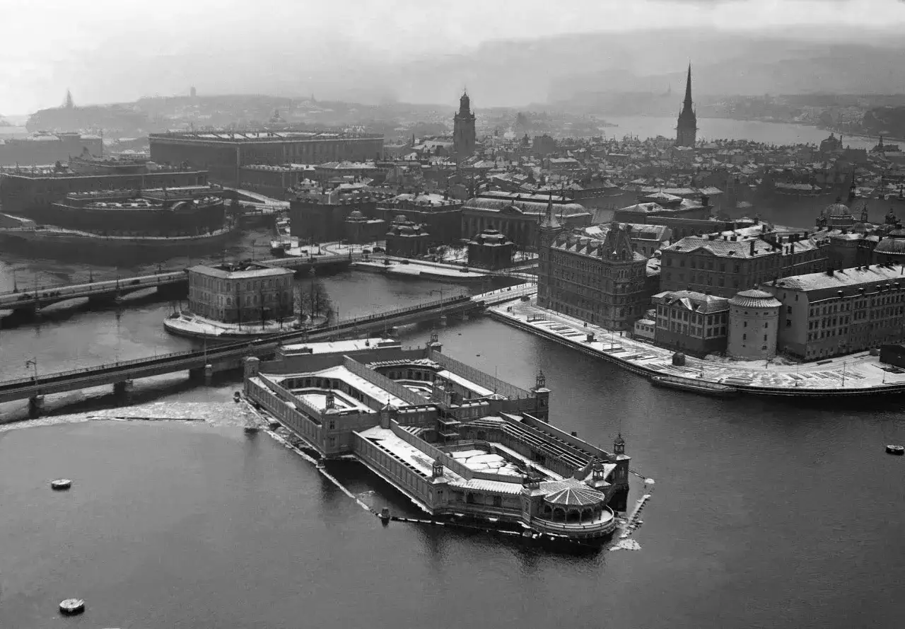 Vintervy över Stockholm med ett imponerande kallbadhus i förgrunden. Snö täcker taken och is flyter i vattnet.