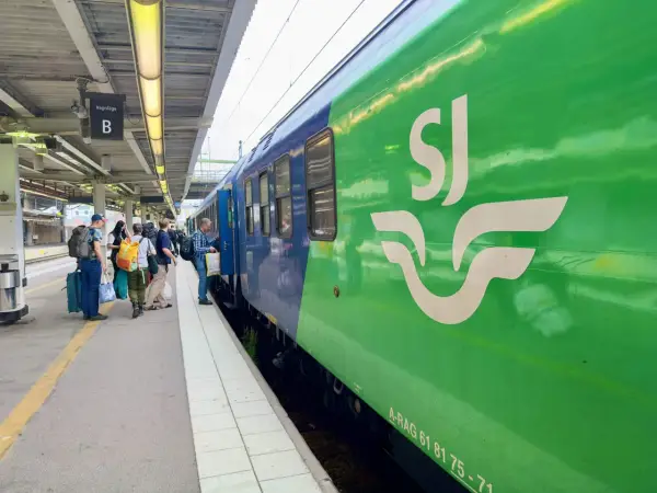 Folk väntar på ett grönt och blått tåg med SJ-loggan på perrongen. Kanske på väg till London?
